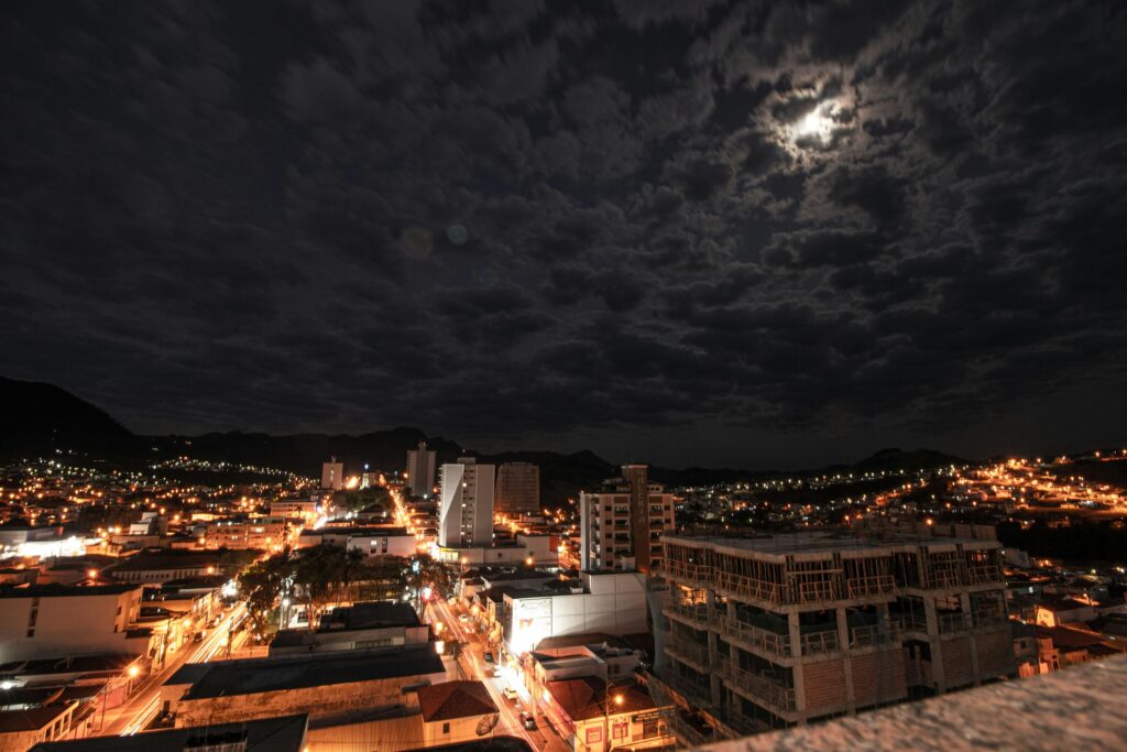 Aerial view of a vibrant cityscape illuminated by lights under a cloudy, moonlit night sky.
