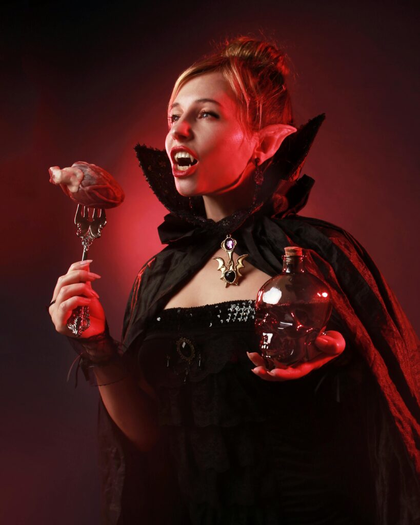 Gothic vampire woman in spooky Halloween costume with a demon-like cape and red lighting.