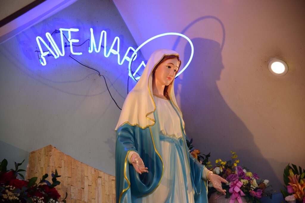 Elegant statue of Virgin Mary under a neon 'Ave Maria' sign with floral decor.